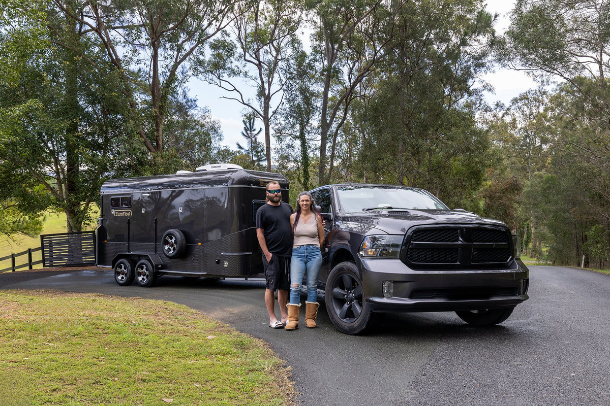 Couple standing beside their black ute and black EuroFloat float, hitched up on a tree-lined driveway