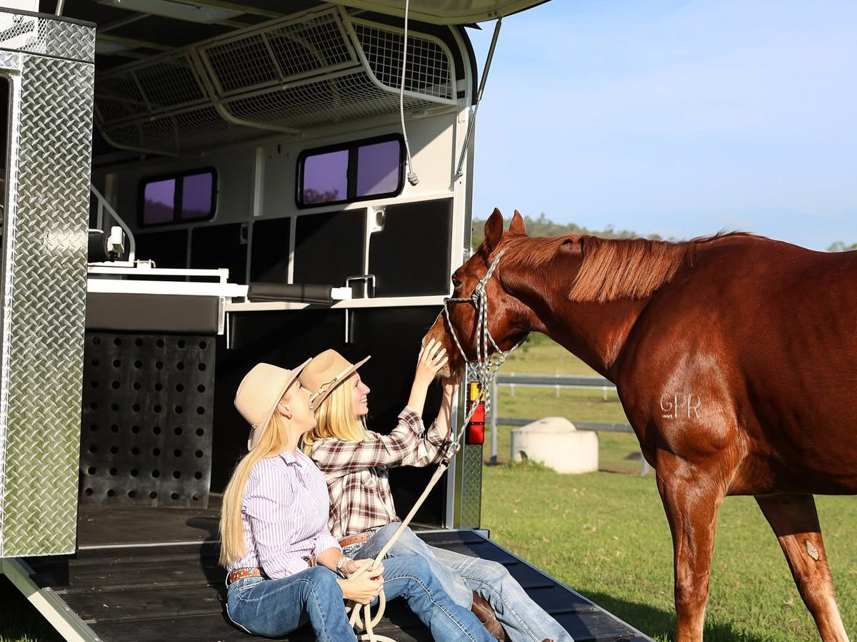Two women in hats sitting on the ramp of a float, patting a chestnut horse